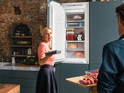 Sieviete, kas stāv pie atvērta ledusskapja un skatās uz vīrieti, kurš rokās tur virtuves dēli; uz dēļa atrodas zemenes.