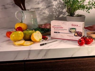 A meatprobe box rests on a marble countertop with colorful vegetables and herbs.