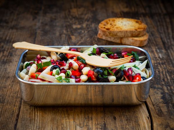 Teglia con ceci, olive nere, melograno, peperoni rossi e scaglie di formaggio, servita con posate in legno e fette di pane