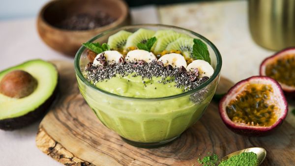 Tagliere con smoothie bowl verde guarnita con kiwi, banana e semi di chia, affiancata da avocado e frutto della passione