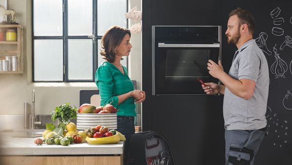 Een monteur staat in een keuken en opent met één hand een oven terwijl hij met de klant praat.