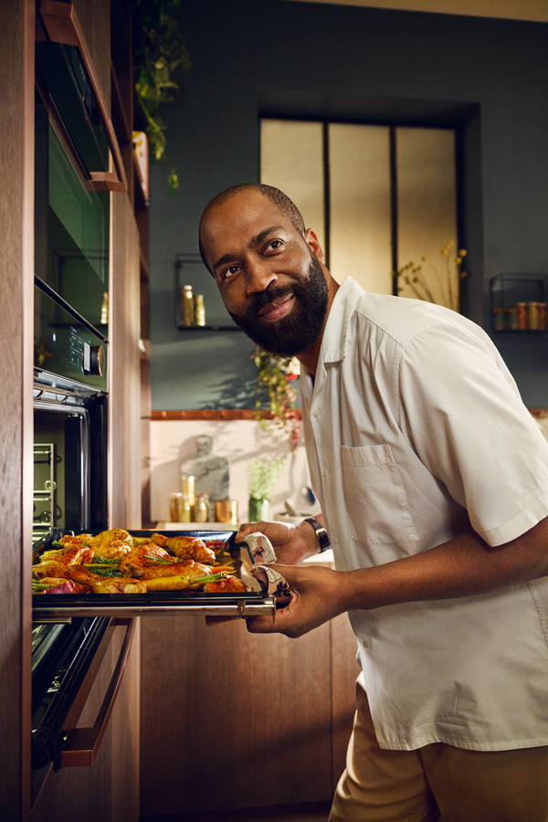 Eine Person in einem weißen Hemd zieht ein Tablett mit gebratenem Huhn und Gemüse aus einem Ofen in einer stilvollen Küche.