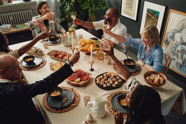 Gruppo di amici che brinda seduto attorno a una tavola apparecchiata con tovaglia chiara, piatti scuri, candele accese e pietanze