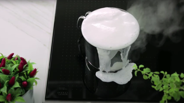 Pentola traboccante di latte schiumoso su superficie di cottura, con vapore, erbe verdi e piante rosse in primo piano. 