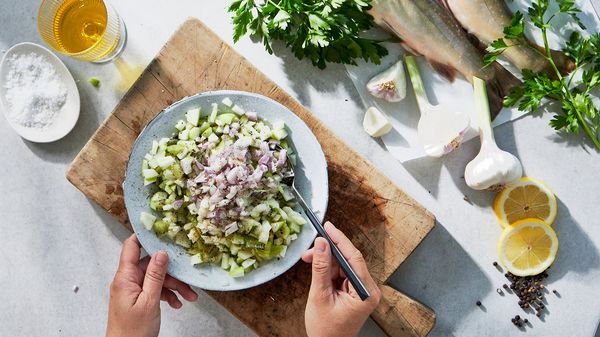 A person mixing finely diced shallots, kiwis and apples in a plate on a table beneath some other ingredients.