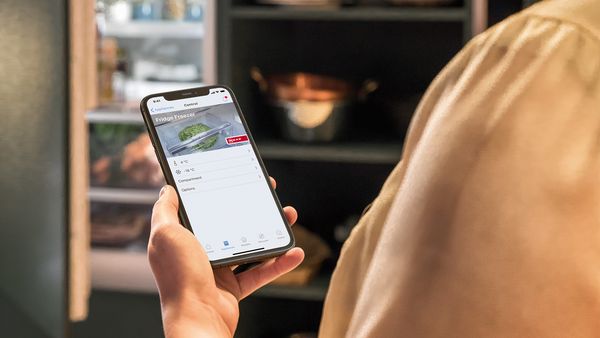 Woman holds smartphone in front of fridge, Home connect app is open