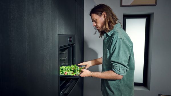 Човек поставя тава със зелени зеленчуци в една отворена фурна в черна кухня