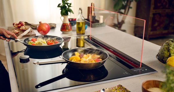 Vue latérale de deux poêles avec des aliments grésillant sur une table de cuisson avec hotte en verre intégrée en haut.