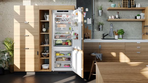 Nyitott beépíthető kombinált hűtőkészülék egy világos fából készült konyhában, nappali fénnyel megvilágítva