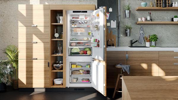 An open built-in fridge freezer inside a light-wood kitchen illuminated by daylight