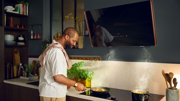 Een man staat te koken met een pan op een flex design kookplaat vóór een hellende afzuigkap gemonteerd aan een groene wand