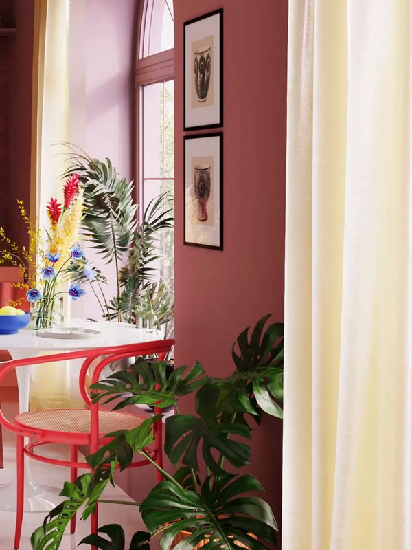 Petite table blanche et chaise rouge devant une grande fenêtre garnie de fleurs colorées. Il y a deux tableaux sur le mur de couleur rouge et une autre plante sur le sol.