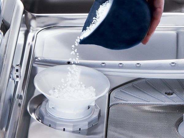 Mano che versa il sale nel serbatoio della lavastoviglie usando un imbuto bianco 