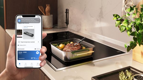 Kött i en metallgryta på en NEFF-häll. I bildens främre del syns en persons hand som håller i en smartphone med Home Connect-appen öppnad.