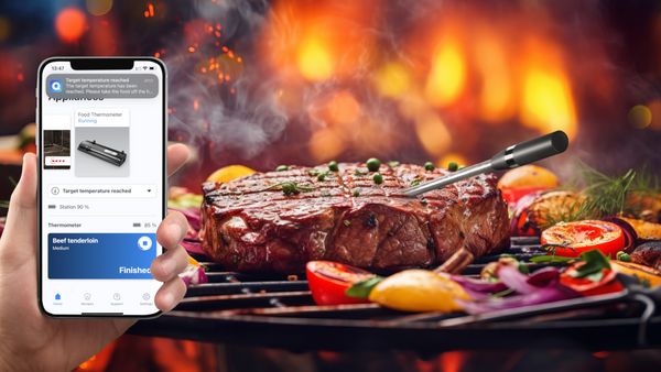 Kött, upphettat på en grill med en trådlös köttsond som sticker in. I bildens främre del syns en persons hand som håller i en smartphone med Home Connect-appen öppnad.