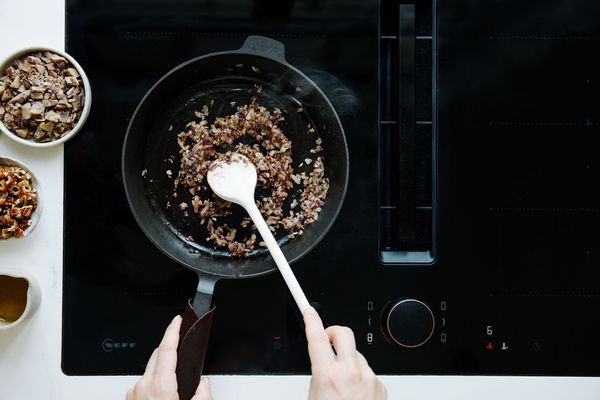 Een persoon roert gesauteerde uien in een zwarte koekenpan op een inductiekookplaat, omringd door schalen met ingrediënten. 