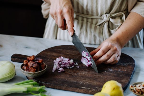 Iemand snijdt rode uien op een houten snijplank, omringd door dadels, groene uien, een citroen en andere ingrediënten. 