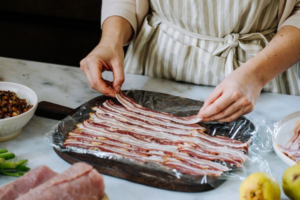 Een persoon in een gestreept schort schikt reepjes bacon op een dienblad bekleed met plasticfolie. 
