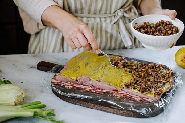 Iemand smeert mosterd op een plat stuk vlees met daarop een notenmengsel en bereidt een gastronomisch gerecht  
