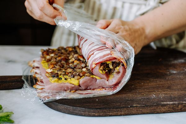 Een persoon pakt een opgerolde schaal met spek, noten en gele vulling uit plasticfolie op een houten snijplank. 