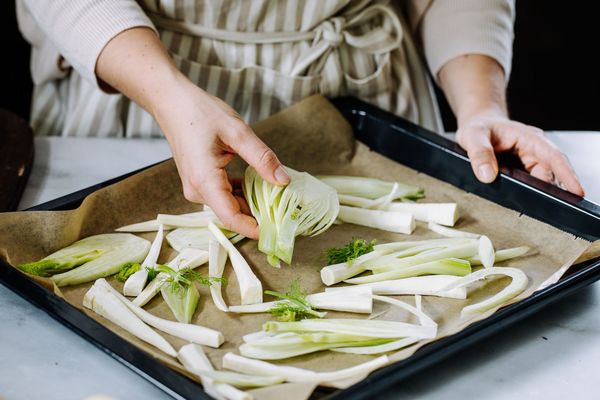 Een persoon met een gestreept schort legt gesneden venkel op een beklede bakplaat, omringd door meer gesneden groenten. 