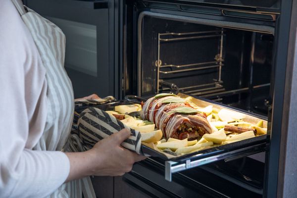 Een persoon in een gestreept schort trekt een bakplaat gevuld met gevuld vlees en aardappelen uit een moderne oven. 