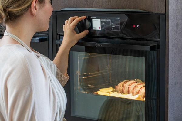 Een persoon regelt de temperatuur van een moderne oven, met een bakplaat met geroosterd vlees en groenten zichtbaar door de glazen deur. 