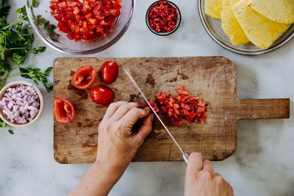 Een hand snijdt tomaten op een houten snijplank, omringd door in blokjes gesneden groenten, kruiden en ananasschijven. 
