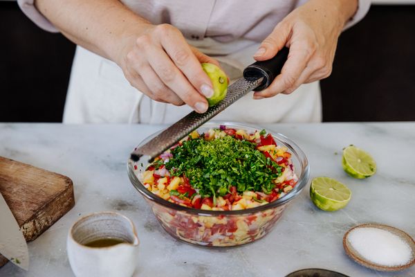 Een persoon schilt een limoen boven een salade van fruit en kruiden in een glazen kom, met keukengerei. 
