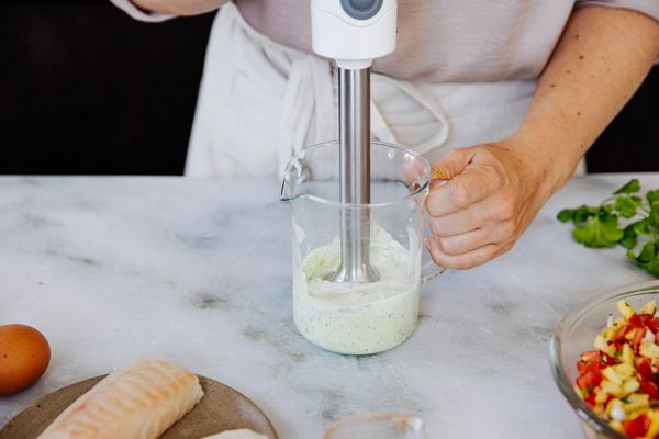 Iemand die met een staafmixer een groene saus mengt in een glazen maatbeker, omringd door verse ingrediënten en rauwe vis. 