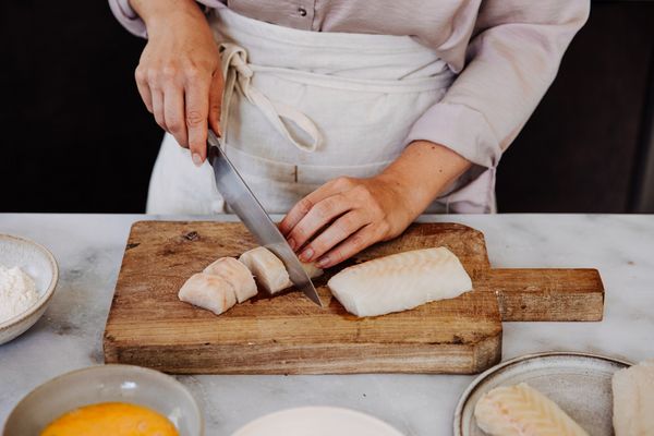 Een persoon snijdt visfilets op een houten snijplank, met kommen bloem en ei in de buurt, in een schone keukenomgeving. 