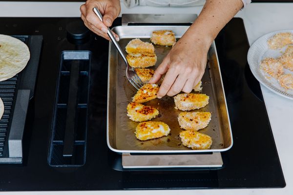 Een hand tilt gouden, gepaneerde stukjes kip van een bakplaat op een modern fornuis, met tortilla's en meer eten. 