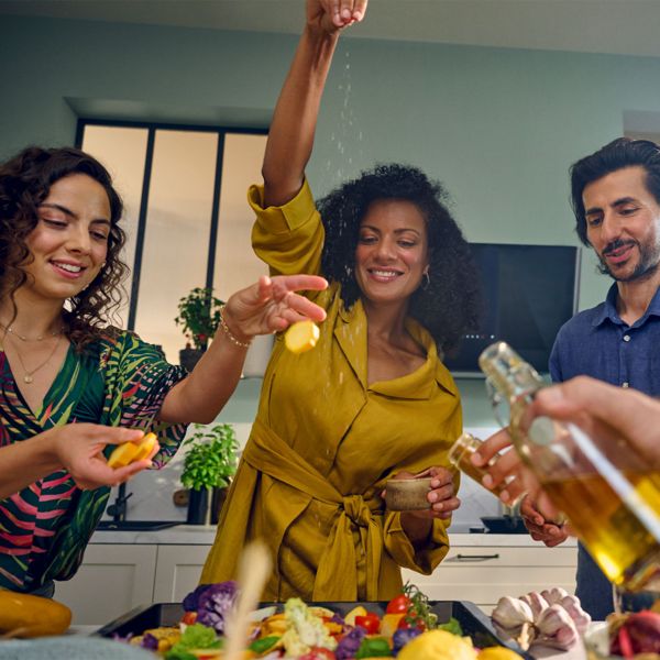 Three friends cooking tray of vegetables, hand adding olive oil