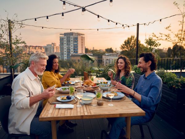 Amici che cenano all&rsquo;aperto su una terrazza, seduti a un tavolo in legno apparecchiato con piatti, bicchieri e lucine sospese