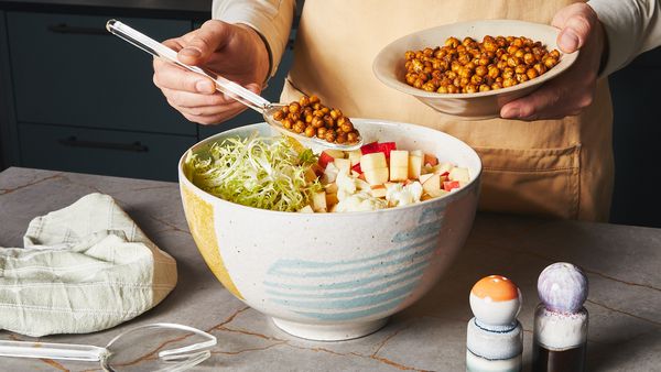 Spooning in air fried chickpeas into a large bowl filled with cut up cauliflower, salad and apple