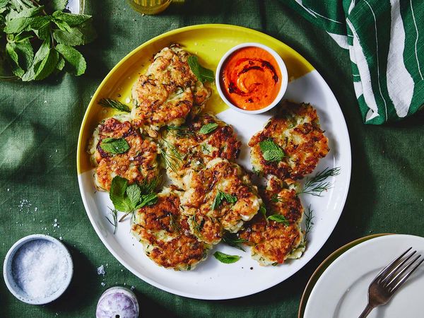 A yellow and white plate with zucchini fritters and red dip on a green table cloth surrounded by colorful dinner table dishes, like a fork, a napkin, plates and a glas filled with water.