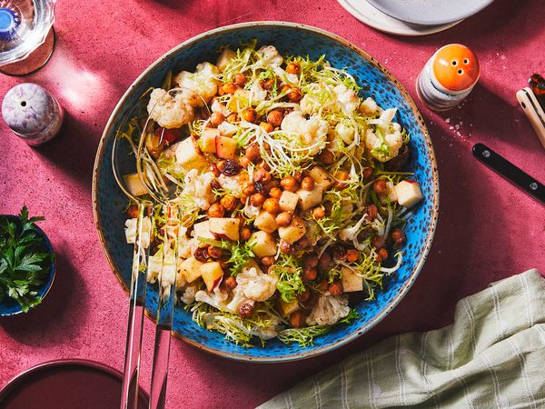 A big blue bowl filled with cauliflower salad surrounded by colorful dinner table dishes, like a fork, a napkin, plates and a glas filled with water.