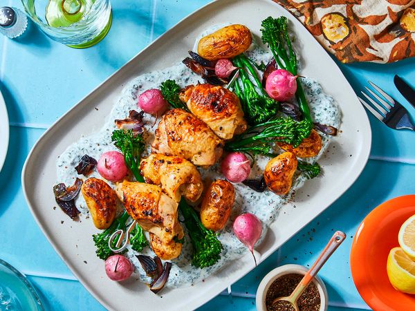 A plate of air fried vegetables and chicken breasts on a herby yogurt dip on a blue underground surrounded by colorful dinner table dishes, like a fork, a napkin, plates and a glas filled with water. 