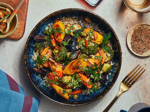 A blue and black ceramic bowl filled with tri colored quinoa which is topped with air fried vegetables is standing in the middle of a table and surrounded by colorful dinner table dishes, like a fork, a napkin, plates and a glas filled with water.