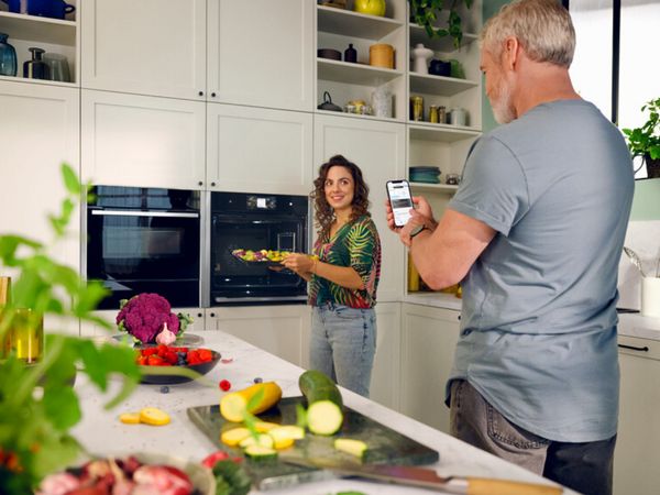 In a kitchen, a man and woman are standing side by side, looking at a phone while discussing something.