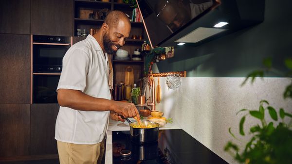 Een man kookt op een inductiekookplaat in een moderne keuken, omringd door houten keukenaccessoires en kruiden, met inbouwovens op de achtergrond
