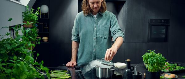 Individual handling a pot in a stylish kitchen, surrounded by fresh herbs and kitchen accessories. 