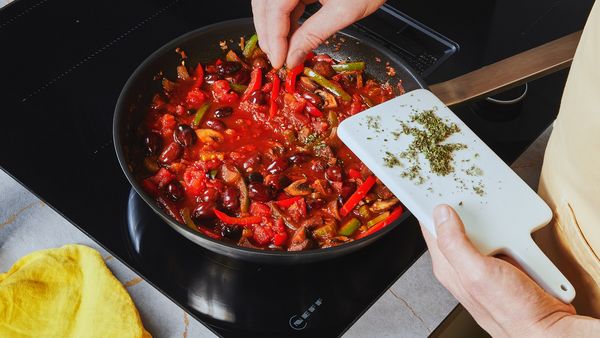 Giving finely cut herbs into a black pot filled with tomato vegetable sauce on the stovetop.