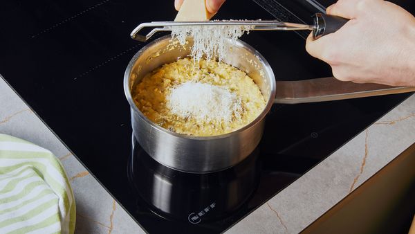 Parmigiano grattugiato aggiunto alla polenta in una pentola argentata su un piano cottura.