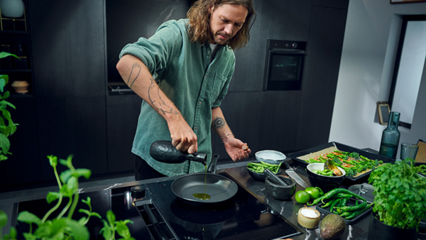 A man prepares a meal in a kitchen, surrounded by fresh vegetables and using a venting hob for cooking.  