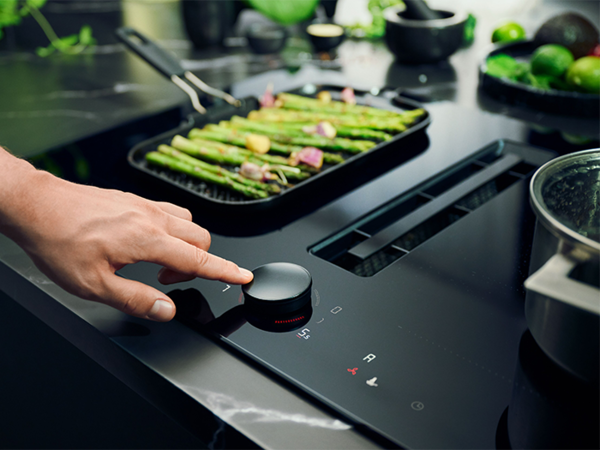 A person touches a venting hob stove top while handling a pan of food, demonstrating cooking techniques.