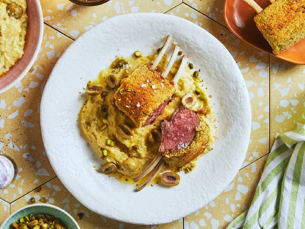 Carré di agnello in crosta di pistacchi con polenta su un piatto bianco posato su piastrelle gialle di ceramica e circondato da altre stoviglie.