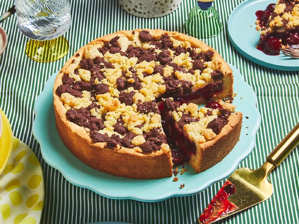 Ein Streuselkuchen mit Sauerkirschen auf einem hellblauen Tortenständer steht auf einer grünen Tischdecke, umgeben von anderem Geschirr.