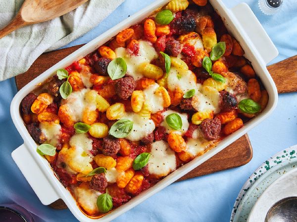 A white baking dish filled gnocchi and sausage in a tomato sauce sits on a light blue tablecloth and is surrounded by other tableware.