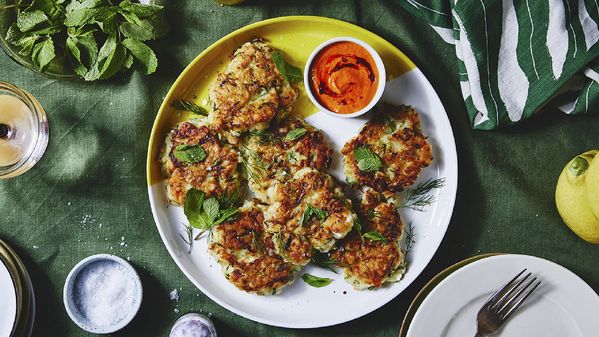 Ein gelb-weißer Teller mit Zucchini-Puffern und rotem Dip auf einer grünen Tischdecke, daneben farbenfrohe Essgedecke, bestehend aus Gabel, Serviette, Tellern und einem Glas Wasser.